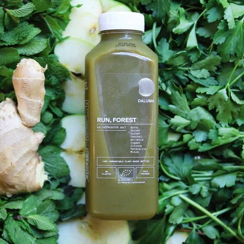 Eine Flasche GRÜNE SAFTKUR - 3 TAGE von DALUMA steht auf Kräutern, Ingwer und Apfelscheiben und präsentiert den reinen, biologisch kalt gepressten Saft für einen natürlichen 3-Tage-Entgiftungsschub.
