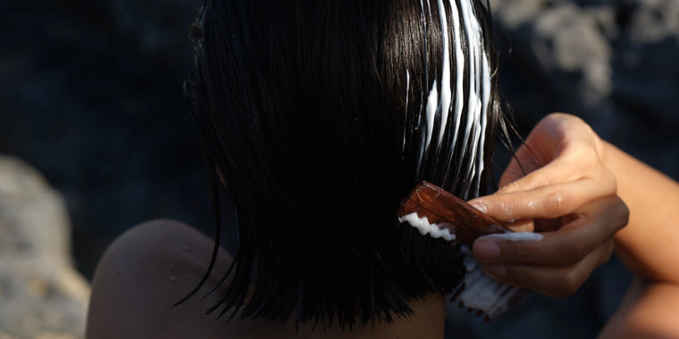 Eine Person trägt mit einem braunen Kamm weiße Pflegespülung auf nasses, dunkles Haar auf, vor einem dunklen, unscharfen Hintergrund.