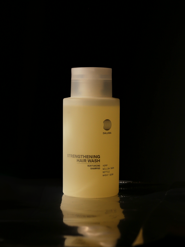 Eine durchsichtige gelbe Flasche mit der Aufschrift Dalluha Strengthening Hair Wash steht aufrecht vor einem schwarzen Hintergrund. Der Text beschreibt das Produkt als nährendes Shampoo mit Hanf, wildem Salbei, Brennnessel und Weizenkeimen.