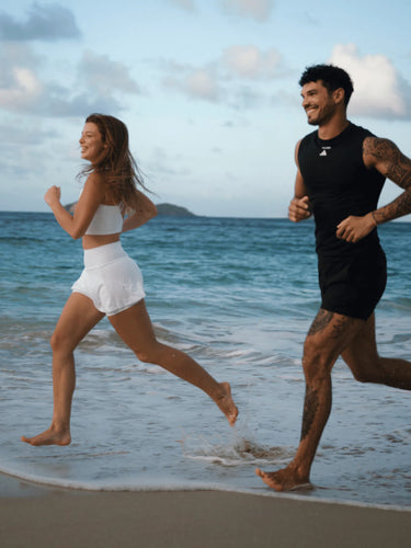 Eine Frau und ein Mann joggen barfuß am Strand entlang, lächeln und genießen die freie Natur. Im Hintergrund sind die Wellen des Ozeans und eine entfernte Insel bei teilweise bewölktem Himmel zu sehen.