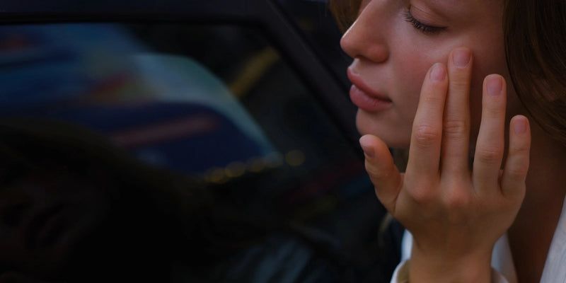 Eine Person mit geschlossenen Augen berührt mit der Hand sanft ihre Wange, wirkt nachdenklich oder in sich gekehrt, neben einem Fenster mit verschwommenen Spiegelungen im Hintergrund.