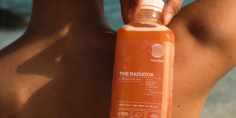Eine Person hält eine Flasche DALUMA The Radiator Saft in der Nähe ihrer nackten Schulter, wobei der Strand und das Meer im Hintergrund verschwimmen. Auf dem Etikett des orangefarbenen Getränks sind Zutaten wie Aloe Vera und Kurkuma aufgeführt.