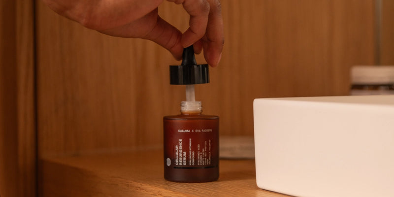 Eine Hand hält einen Tropfer über einer offenen Braunglasflasche mit der Aufschrift Serum, die auf einem Holzregal neben einem weißen rechteckigen Gegenstand steht.