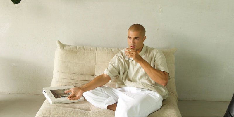 Ein Mann in einem beigen Hemd und einer weißen Hose sitzt im Schneidersitz auf einem cremefarbenen Sofa, in der einen Hand ein Getränk, in der anderen ein Buch, vor einer minimalistischen hellen Wand.