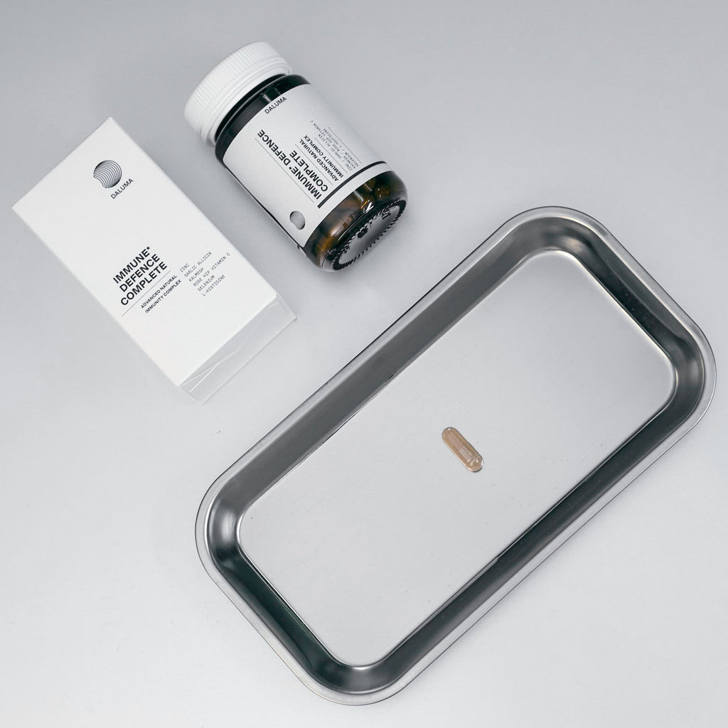 Eine einzelne Kapsel auf einem Metalltablett neben einer weißen DALUMA IMMUN DEFENCE COMPLETE KOMPLEX-Schachtel und einem Pillenfläschchen, die den Knoblauchextrakt zur Stärkung des Immunsystems hervorheben, vor einem hellen Hintergrund.