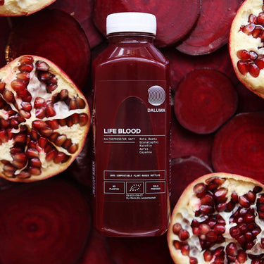 Eine Packung DALUMA LIFE BLOOD POWER SAFT 6 PACK, mit Kaltgepresster Saft, ist umgeben von Granatapfel- und Rübenschnitzen. Das Etikett hebt Zutaten wie rote Beete Saft und Granatapfelsaft hervor und betont die umweltfreundliche Verpackung.