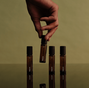 Eine Hand hält eine bernsteinfarbene Rollflasche der NULLPUNKT COMPLETE COLLECTION von Nullpunkt über einer reflektierenden Oberfläche. Drei passende Flaschen und minimalistische Etiketten stehen vor einem Hintergrund in gedämpftem Grün für einen eleganten, natürlichen Look.