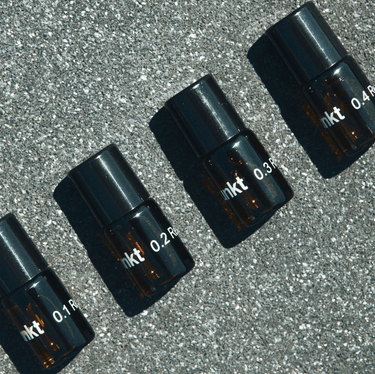 Vier kleine Braunglasflaschen mit schwarzen Deckeln, die mit "0.1", "0.2", "0.3" und "0.4" beschriftet sind, aus dem Nullpunkt NULLPUNKT DISCOVERY SET sind diagonal auf einer gesprenkelten grauen Fläche angeordnet und werfen Schatten nach rechts.