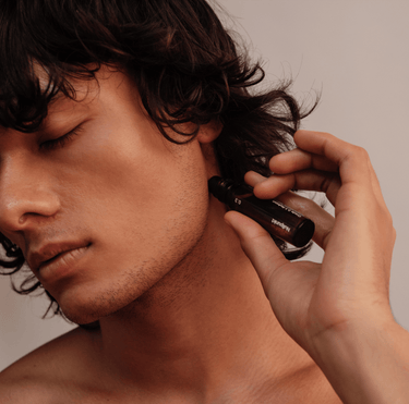 Eine Person mit gewelltem dunklem Haar und geschlossenen Augen trägt Nullpunkt Grade 0.1 Relieve Öl aus einer kleinen braunen Flasche auf ihren Hals auf und fängt einen ruhigen, beruhigenden Moment mit diesem beruhigenden Nullpunkt Produkt ein.