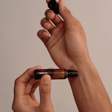 Eine Person hält einen kleinen Roll-on-Flakon aus Braunglas mit der Aufschrift NULLPUNKT GRADE 0.3 REFOCUS von Nullpunkt in der Hand und schraubt den schwarzen Verschluss mit einer Hand ab, während sie den Flakon vor einem minimalistischen beigen Hintergrund hält.