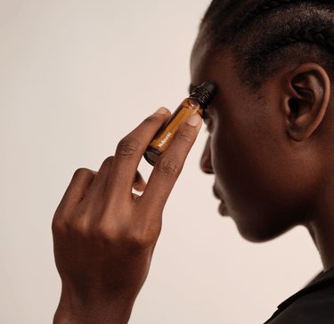 Eine Person mit geflochtenen Haaren trägt die Rollerflasche Nullpunkt NULLPUNKT GRADE 0.4 REFUEL sanft an der Schläfe auf und hält sie vor einem neutralen Hintergrund dicht an ihr Gesicht.