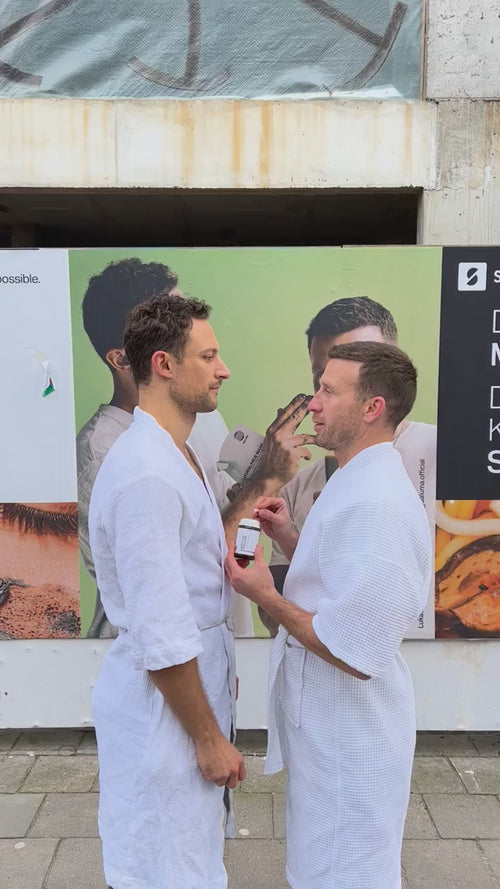 Zwei Männer in weißen Bademänteln stehen sich auf einem Bürgersteig gegenüber, wobei einer von ihnen eine kleine Schachtel in der Hand hält. Hinter ihnen befindet sich eine Wand mit bunten Plakaten, darunter Bilder von Lebensmitteln und Menschen.