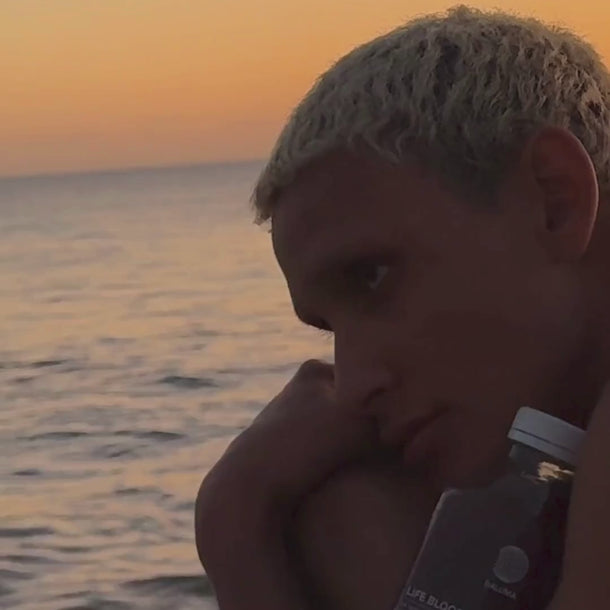 Eine Person mit kurzem, hellem Haar sitzt bei Sonnenuntergang am ruhigen Meer und hält eine klare Flasche Daluma BETTER GUT SAFTKUR 3 TAGE in der Hand, während der Himmel im Hintergrund orange leuchtet.