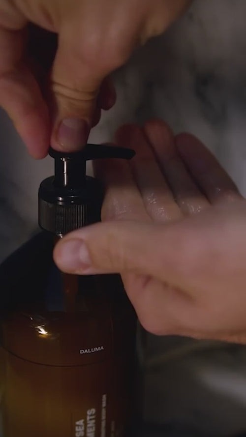 Eine Nahaufnahme zeigt Hände, die auf die Pumpe einer braunen Daluma BODY CARE DUO-Flasche drücken, um Flüssigseife auf eine offene Handfläche abzugeben, mit einem unscharfen Hintergrund.