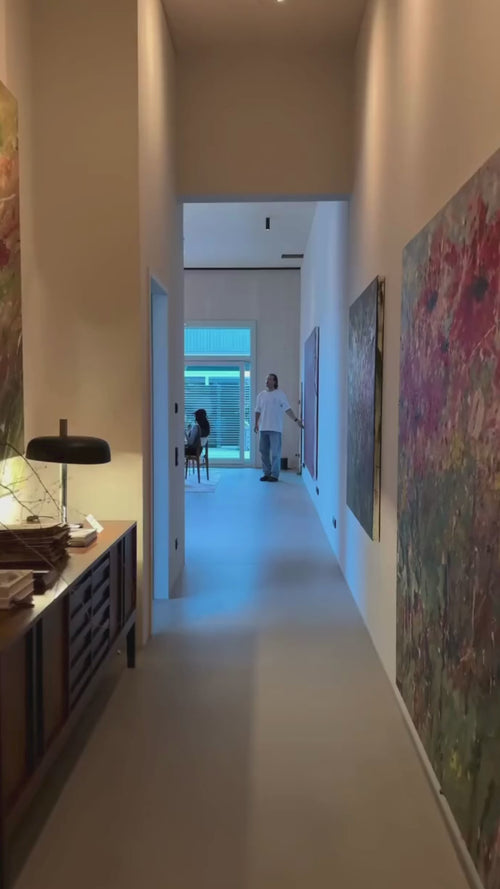 Ein Flur mit bunten abstrakten Gemälden an den Wänden führt in einen hellen Raum, in dem eine Person neben einer sitzenden Person an großen Fenstern steht. Auf der linken Seite steht ein Beistelltisch mit einer Lampe und Dekoration.