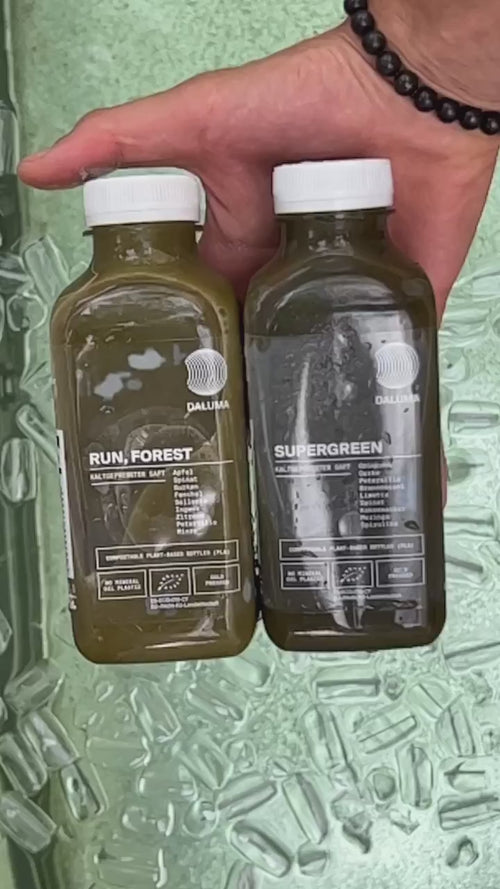 Eine Hand hält zwei grüne Daluma-Saftflaschen aus dem "GRÜNE SAFTKUR - 5 TAGE"-Programm mit weißen Verschlüssen und Etiketten mit weißer Schrift und Grafik über verstreuten Eiswürfeln.