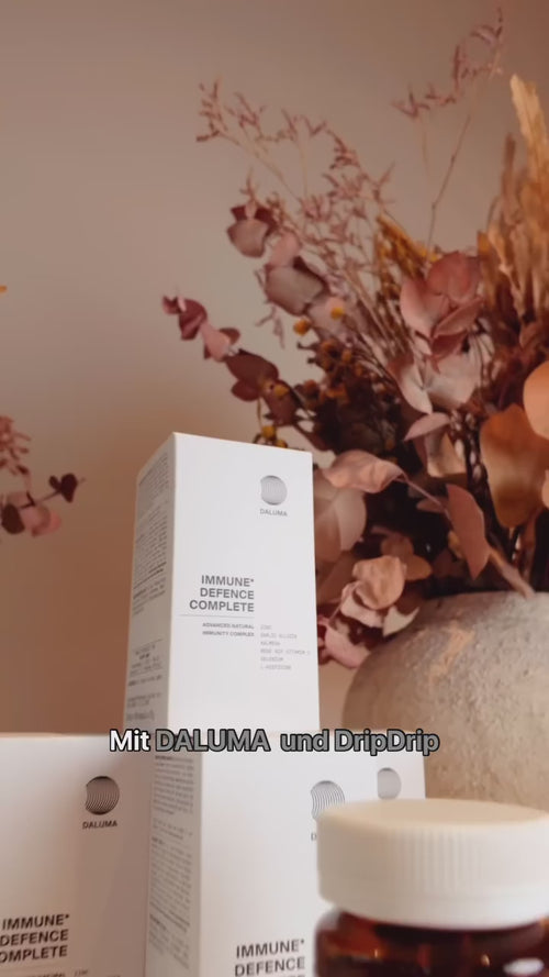 Weiße Schachteln mit DALUMA Immune Defence Complete Nahrungsergänzungsmitteln sind vor getrockneten Blumen gestapelt. Der deutsche Text auf dem Bild lautet: Mit DALUMA und DripDrip.