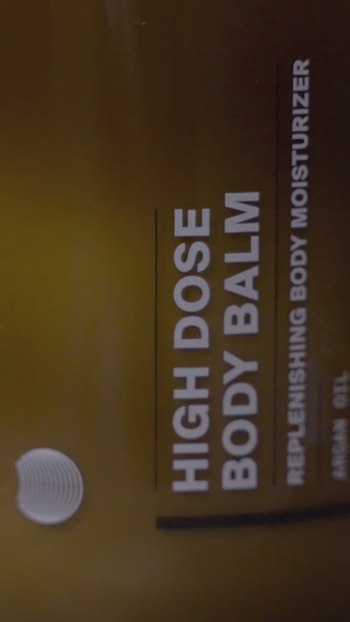 Eine Nahaufnahme einer braunen Flasche mit der Aufschrift HIGH DOSE BODY BALM und REPLENISHING BODY MOISTURIZER in weißer Schrift. Der Hintergrund ist unscharf und konzentriert sich auf den Produktnamen.