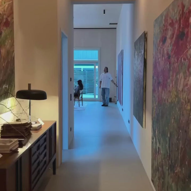 Ein Flur mit großen abstrakten Gemälden führt in einen hellen Raum, in dem ein Mann steht und eine Frau an einem Tisch sitzt. Auf der linken Seite stehen Dalumas HEALTH CLUB PRO MEMBERSHIP-Materialien auf einem Sideboard mit einer Lampe und Büchern.