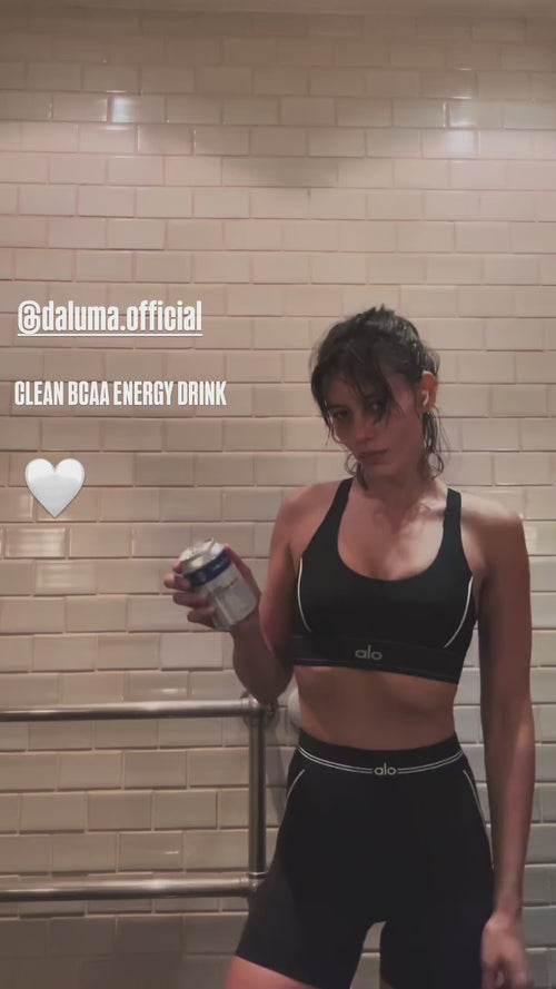 Eine Frau in schwarzem Sport-BH und Shorts steht vor hellbraun gekachelten Wänden und hält eine Dose Daluma CLEAN BCAA ENERGY DRINK. Der Text lautet @daluma.official CLEAN BCAA ENERGY DRINK mit einem weißen Herz-Emoji.