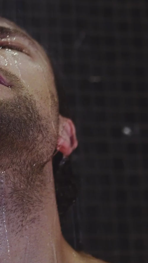 Ein Mann steht mit geschlossenen Augen unter der Dusche, das Wasser rinnt ihm über Gesicht und Hals, auf dunklen Kacheln - und zeigt Dalumas HAIR CARE DUO.