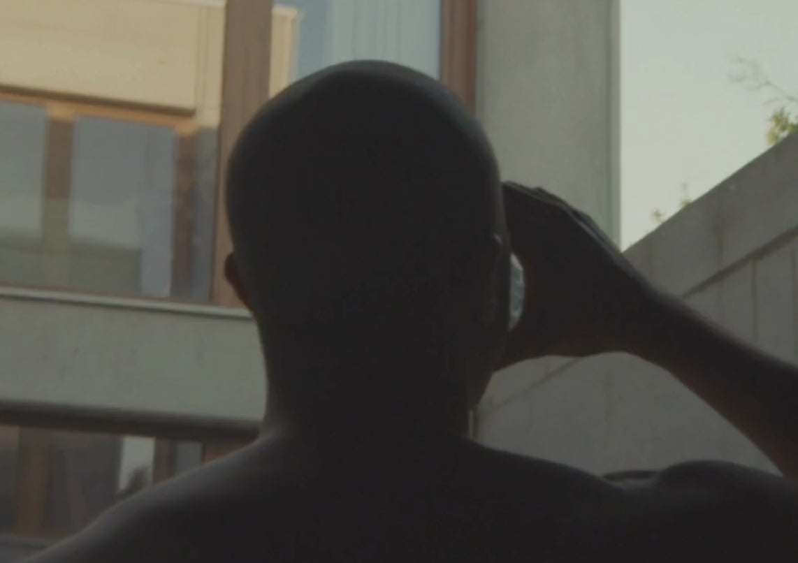 Eine Person mit rasiertem Kopf ist von hinten zu sehen, die ein Glas an den Mund hält und in einem Innenraum in der Nähe eines großen Fensters steht, durch das Sonnenlicht einfällt.