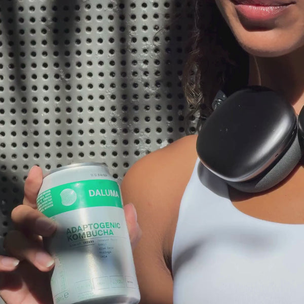 Eine Person in einem weißen Oberteil hält eine Dose Daluma Adaptogenic Kombucha in der Hand. Sie hat schwarze Kopfhörer um den Hals und steht vor einem perforierten Metallhintergrund.