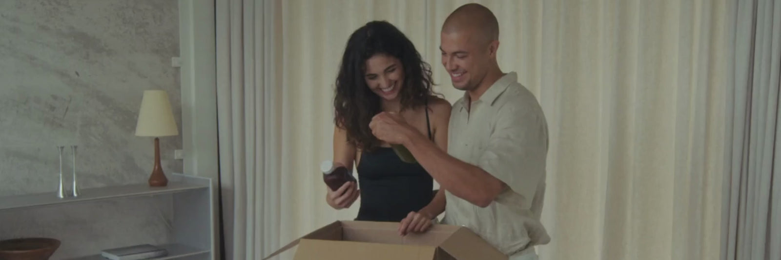Ein lächelndes Paar steht zusammen vor beigen Vorhängen und öffnet einen Karton. Die Frau hält einen kleinen Gegenstand aus der Schachtel, während der Mann zusieht, beide wirken glücklich und aufgeregt.