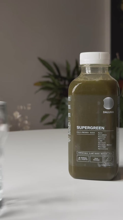 Eine Flasche des Daluma DAILY HEALTH PACKAGE Saftes steht auf einem weißen Tisch, umgeben von Pflanzen und einem unscharfen Hintergrund. Das klare Etikett zeigt die Inhaltsstoffe und hebt die umweltfreundlichen Verpackungsmerkmale hervor.