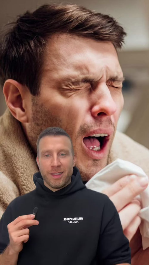 Im Hintergrund ist ein Mann zu sehen, der in ein Taschentuch niest, während ein anderer Mann in einem schwarzen Kapuzenpulli im Vordergrund steht, spricht und ein kleines Mikrofon hält.