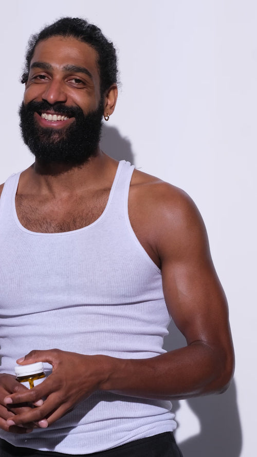Ein lächelnder, bärtiger Mann in einem weißen Tank-Top steht vor einem schlichten, hellen Hintergrund und hält eine Daluma 360 DAILY ESSENTIALS SUPPLEMENT DOUBLE Flasche.