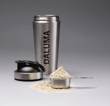Der DALUMA PROTEIN SHAKER EDELSTAHL steht aufrecht mit seinem Deckel daneben; ein Messbecher aus Metall und ein Messlöffel gefüllt mit veganem Proteinpulver zeigen eine nachhaltige Alternative für die tägliche Ernährung.