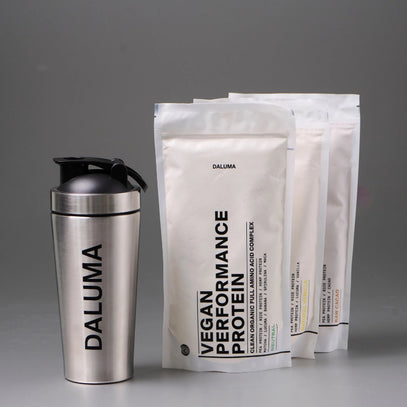 Ein Shaker aus rostfreiem Stahl mit der Aufschrift DALUMA steht neben drei weißen Beuteln mit der Aufschrift VEGAN PERFORMANCE PROTEIN und anderen Produktdetails, alles auf grauem Hintergrund.