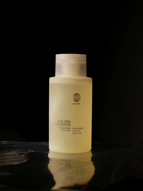 Eine durchsichtige gelbe Flasche mit der Aufschrift "Daluma Raw Sea Elements Regenerating Body Wash" steht vor einem schwarzen Hintergrund. Auf dem Etikett werden Inhaltsstoffe wie Blasentang und Aloe vera erwähnt.