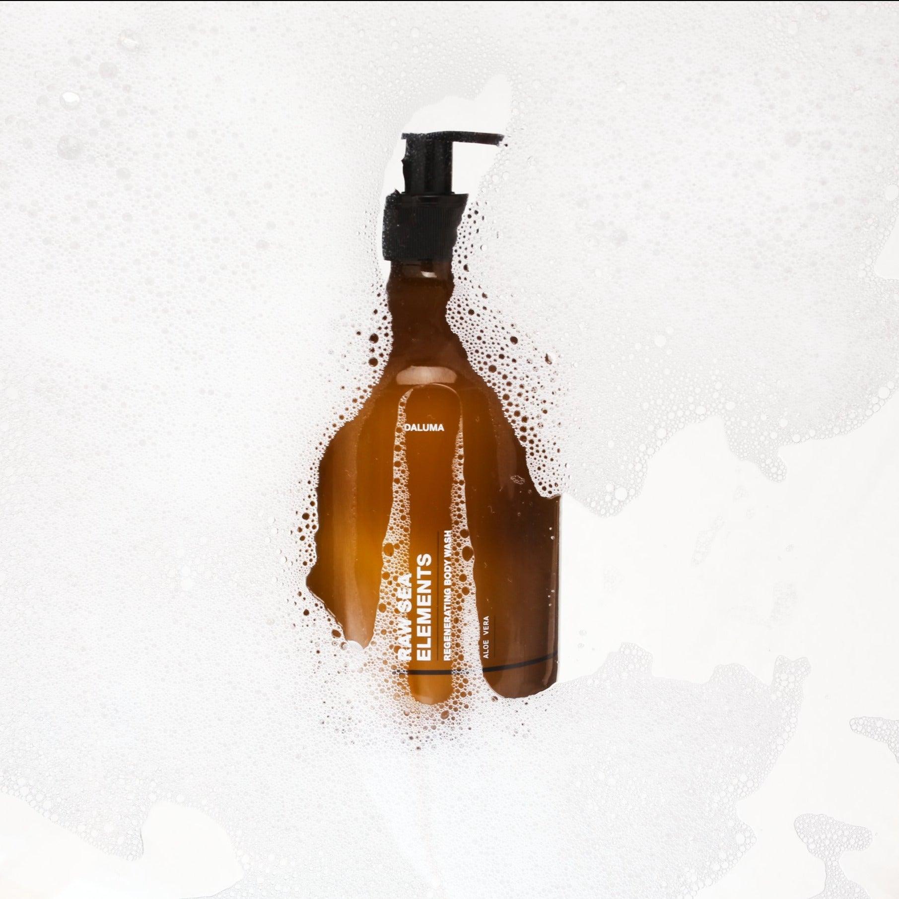 Eine braune Flasche mit einer schwarzen Pumpe und der Aufschrift DALUMA ist teilweise durch die weiße Seifenlauge hindurch zu sehen und weist auf die reinigende Wirkung von RAW SEA ELEMENTS SHOWER GEL hin.