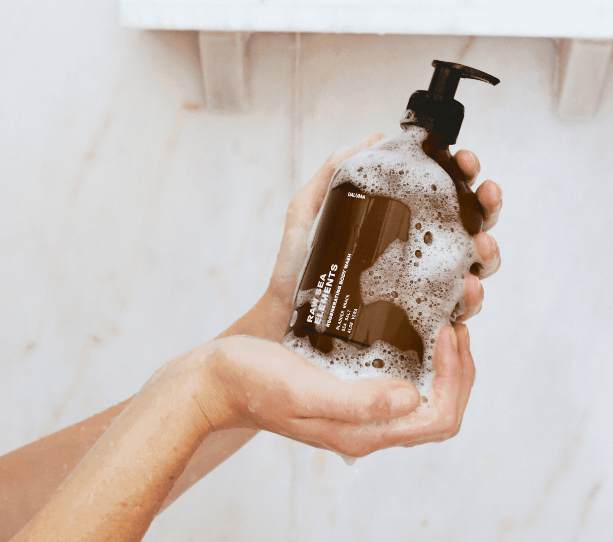 Eine Person hält mit beiden Händen eine braune Daluma-shop BODY CARE DUO Pumpflasche mit schaumiger Seifenlauge vor einer hellen Badezimmerwand und einem weißen Regal im Hintergrund.