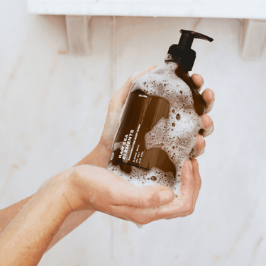 Eine Person hält mit beiden Händen eine Daluma-shop BODY CARE DUO Flasche mit Seifenlauge in einer Dusche vor einer weiß gefliesten Wand und einem Regal im Hintergrund.