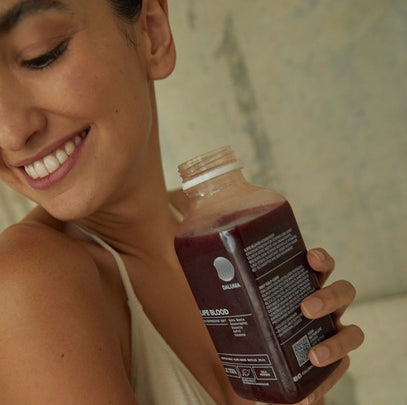 Eine Frau mit dunklem Haar lächelt, während sie eine Flasche dunkelroten Saft mit der Aufschrift Lifeblood in der Hand hält. Sie trägt ein beiges Oberteil und schaut über ihre Schulter.