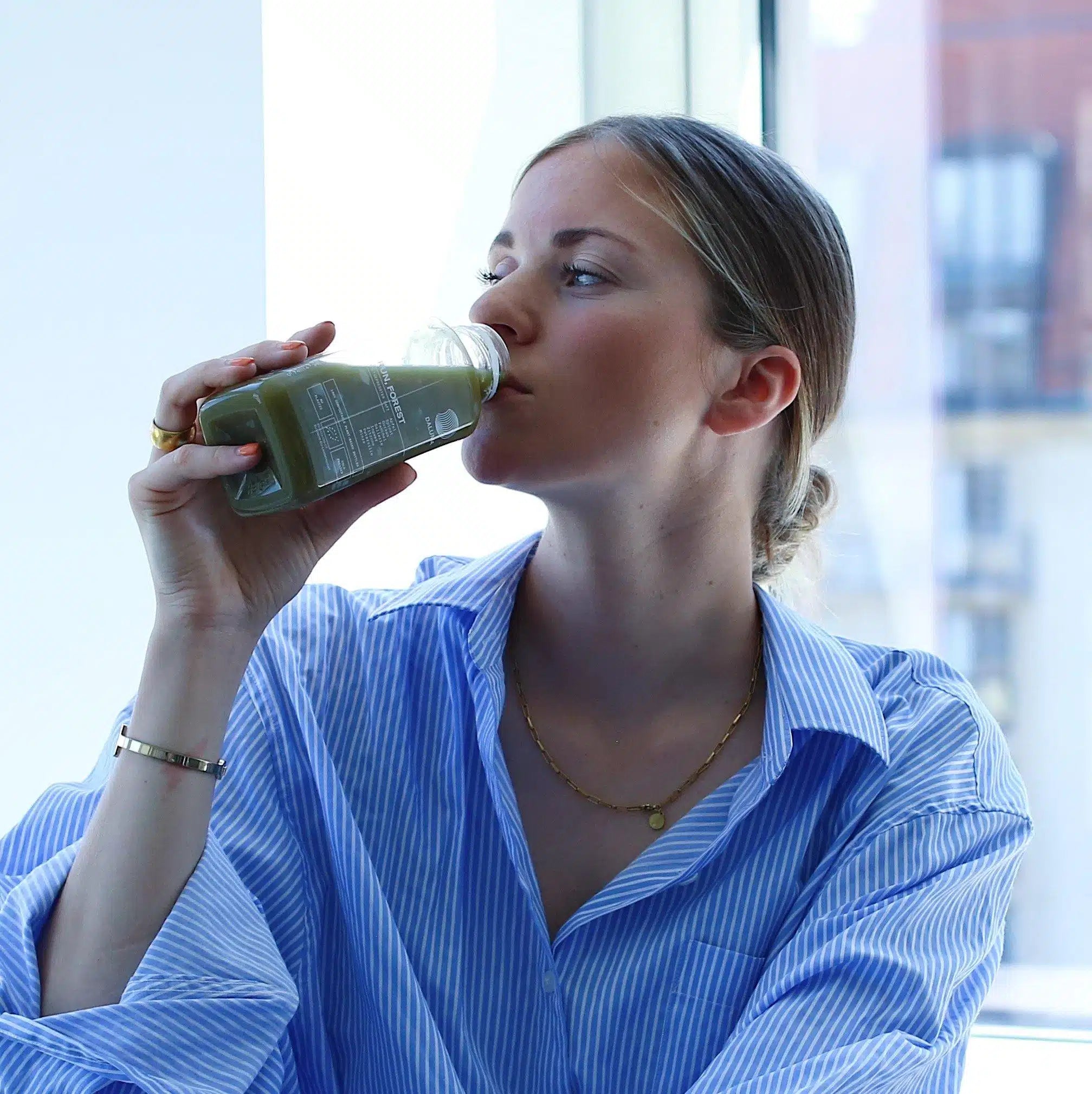 Eine Frau in einem blau gestreiften Hemd schlürft den grünen Smoothie GRÜNE SAFTKUR PLUS - 5 TAGE von DALUMA aus einer Glasflasche an einem Fenster, während die Gebäude der Stadt im Hintergrund verschwimmen - perfekt für Ihre nächste fünftägige Saftkur.