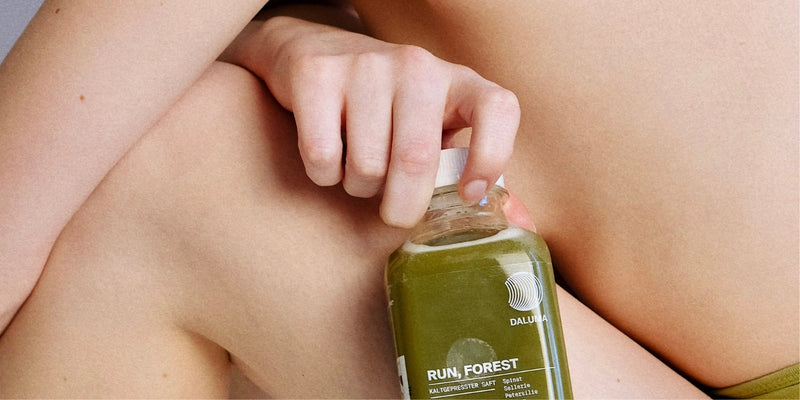 Eine Person mit nackter Haut hält eine durchsichtige Flasche mit grünem Saft mit der Aufschrift Run, Forest by Dalluma in der Hand und stützt sich auf ihr Knie. Das Bild ist eng beschnitten und auf die Flasche und die Hand fokussiert.