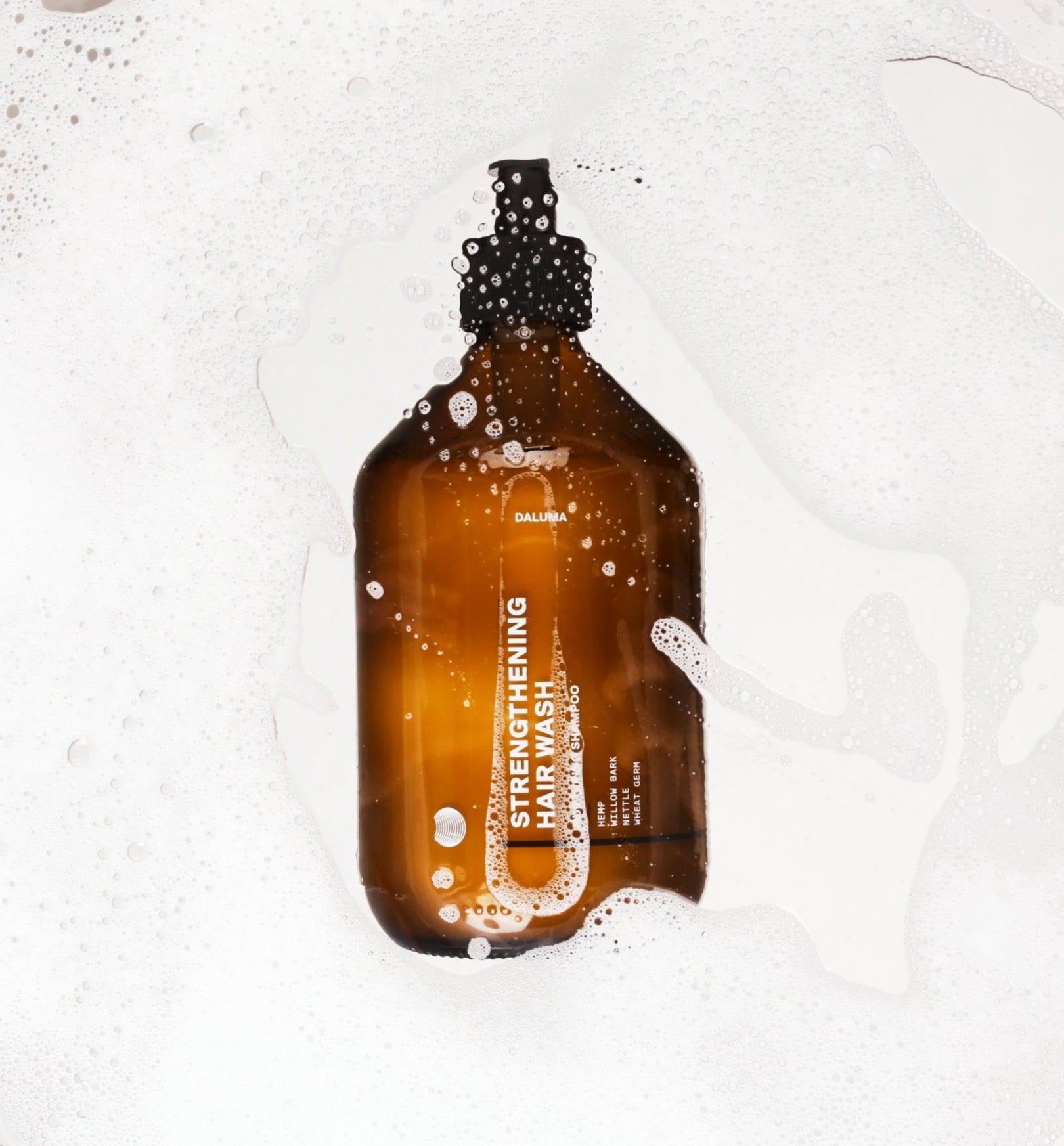 Eine braune DALUMA STRENGTHENING HAIR WASH Flasche mit schwarzer Pumpe liegt in Seifenwasser, spendet intensive Feuchtigkeit und unterstützt das Haarwachstum für kräftiges, gesundes Haar.
