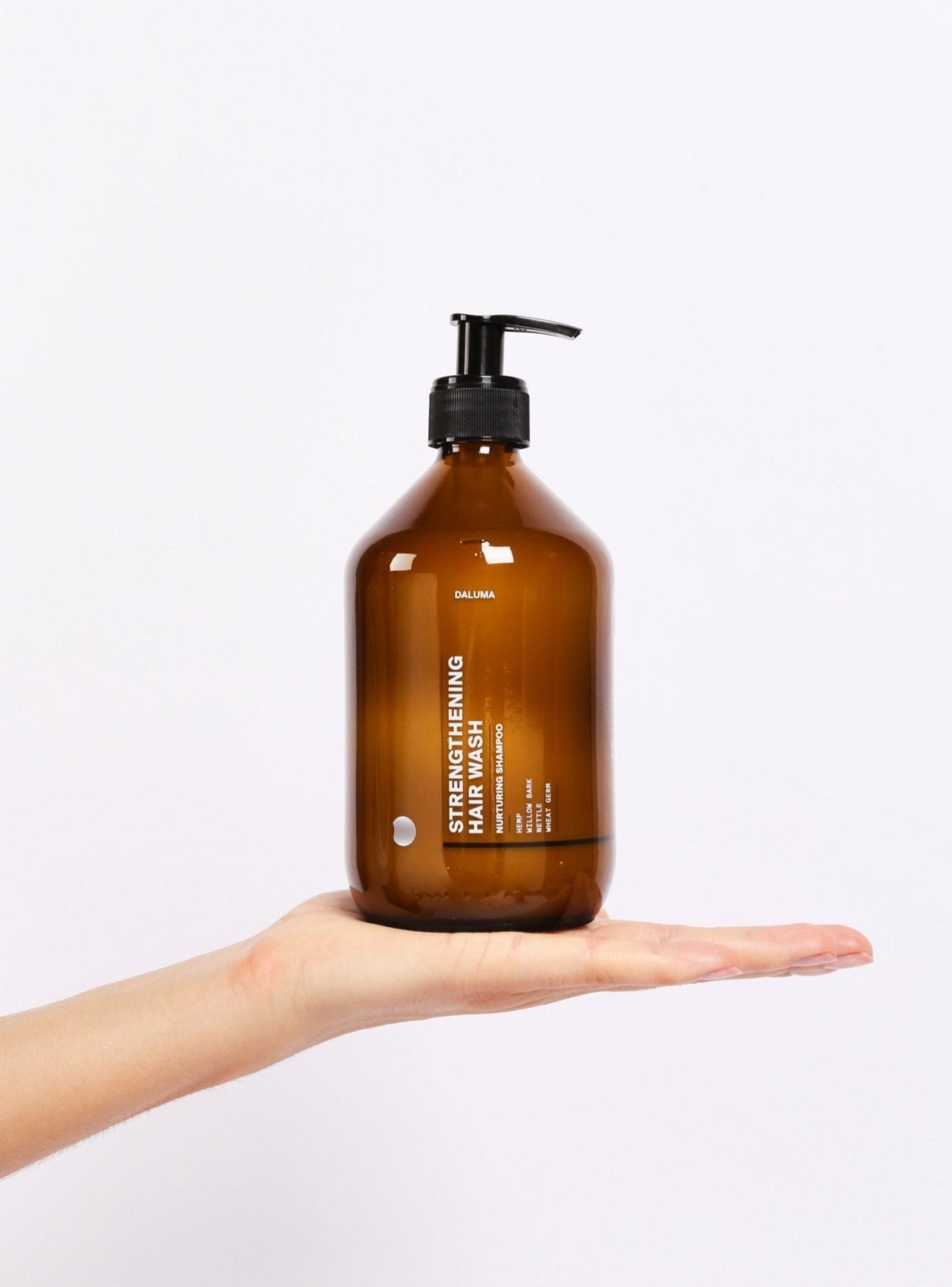 Eine Hand hält eine große bernsteinfarbene Pumpflasche DALUMA STRENGTHENING HAIR WASH, die zur Unterstützung des Haarwachstums entwickelt wurde, vor einem schlichten weißen Hintergrund.