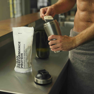 Eine Person mit nacktem Oberkörper und einem Handtuch um die Taille schöpft DALUMA VEGANES PERFORMANCE PROTEIN KAKAO DOUBLE Pulver in eine Shakerflasche auf dem Küchentisch. Der Beutel mit dem Proteinpulver und der Deckel des Shakers sind in der Nähe zu sehen.