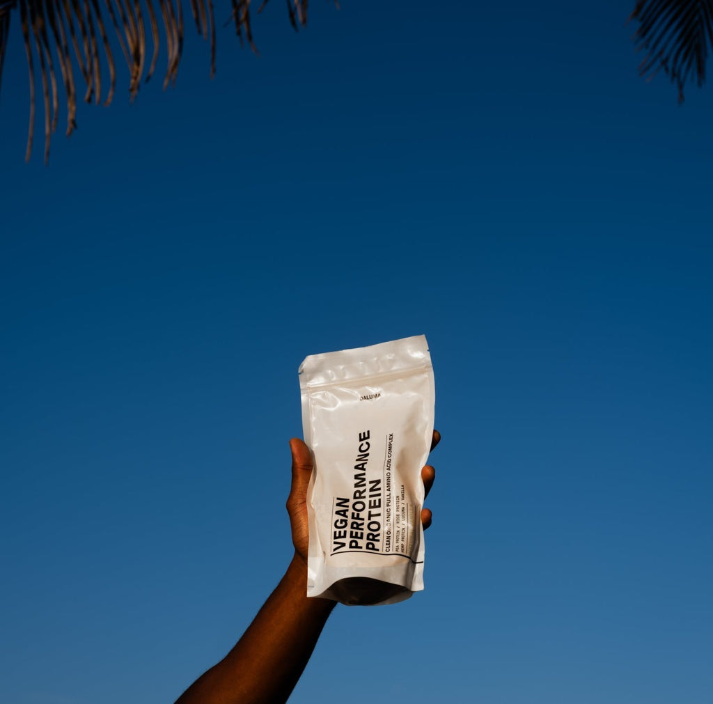 Eine Hand hält einen weißen DALUMA-Beutel mit der Aufschrift VEGANES PERFORMANCE PROTEIN NEUTRAL vor einem klaren blauen Himmel mit Palmenblättern am oberen Bildrand hoch.