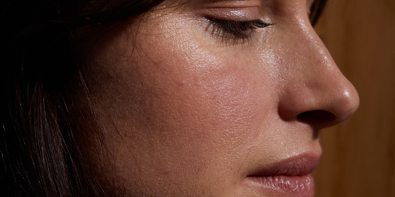 Nahaufnahme des Gesichts einer Person mit geschlossenen Augen, glatter Haut und natürlicher Textur; braunes Haar umrahmt teilweise das Gesicht und ein hölzerner Hintergrund ist sichtbar.